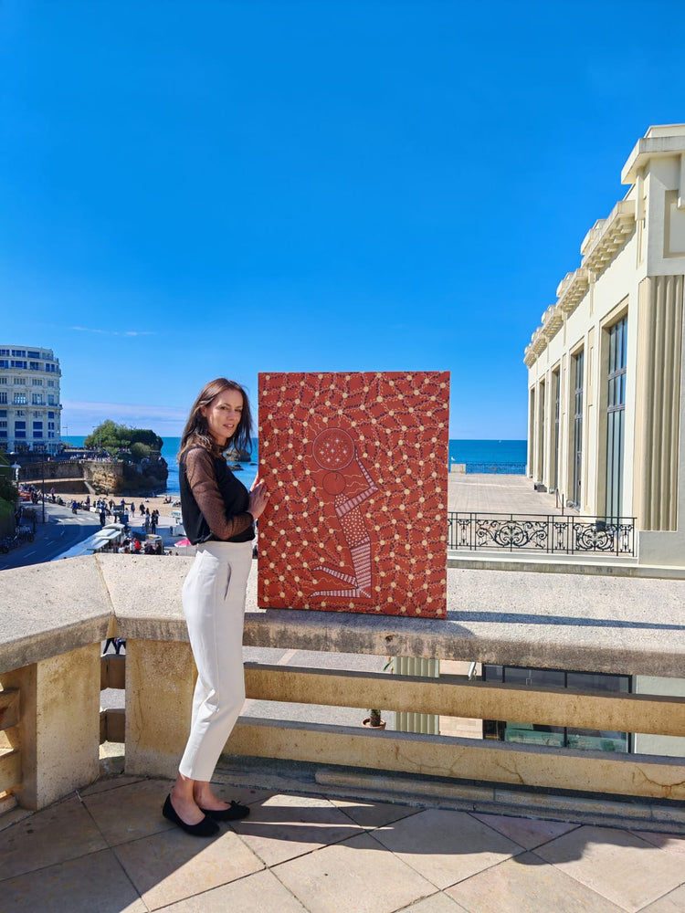 Dorothée Rodriguez-Velar presenting her original artwork, a large abstract red painting, outdoors against a clear blue sky and classical architecture.